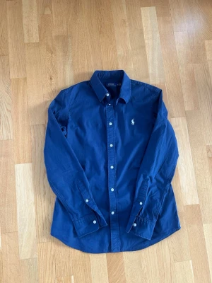 Marinblå skjorta från Polo Ralph Lauren - Klassisk marinblå skjorta från Polo Ralph Lauren med vit broderad logga på bröstet. Skjortan har lång ärm, knappar framtill och vid ärmslut samt en klassisk krage. Tillverkad i bomull för en skön känsla.