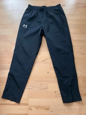 Svarta träningsbyxor Under Armour - Säljer ett par svarta träningsbyxor från Under Armour med resår i midjan och dragkedjor vid bensluten. Byxorna har logga på vänster lår och är tillverkade i ett lätt, snabbtorkande material som passar perfekt till träning.