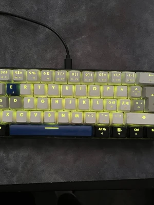 Mekaniskt tangentbord med RGB-belysning - Säljer ett kompakt mekaniskt tangentbord med RGB-belysning som kan ändras mellan olika färger, t.ex. gult och rosa. Tangenterna är vita med svart text och har en blå mellanslagstangent. Tangentbordet ansluts via USB och har nordisk layout med specialtecken.