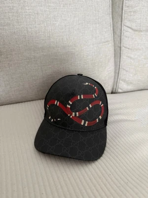 Svart Gucci keps med ormtryck - Säljer en svart keps från Gucci med det klassiska GG-mönstret och en röd-vit orm broderad på framsidan. Kepsen har justerbar rem bak med röd-blå detalj och mesh på sidorna för extra ventilation. Perfekt för dig som vill sticka ut med en lyxig accessoar. ÄKTA!!