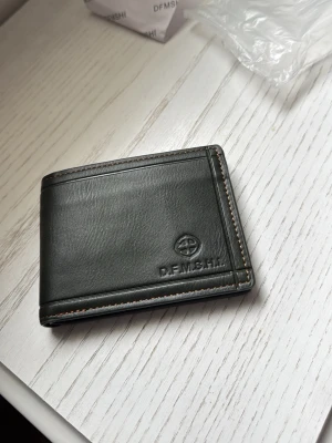 Svart läderplånbok från DFMSHI - Snygg svart plånbok i äkta läder från DFMSHI. Plånboken har bruna detaljer på insidan, flera kortfack och en liten dragkedjeficka för mynt. Klassisk design med diskret logga på framsidan. Perfekt storlek för fickan.