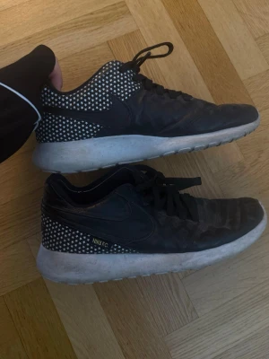 Nike FC svarta sneakers med stjärnor - Nike FC sneakers i svart med vita stjärnor på hälen och sidan. Skorna har en vit, tjock sula och klassisk låg profil. Ovandelen är i textil och syntet med svarta skosnören. Swoosh-logga på sidan och Nike FC-detalj i vitt.