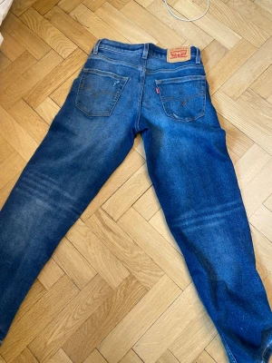 Blå Levi's jeans med raka ben - Säljer ett par klassiska blå Levi's jeans med raka ben och snygga slitningar. Jeansen har två bakfickor, tydliga sömmar och det ikoniska läderpatchen bak i midjan. Perfekta för en avslappnad och tidlös stil.
