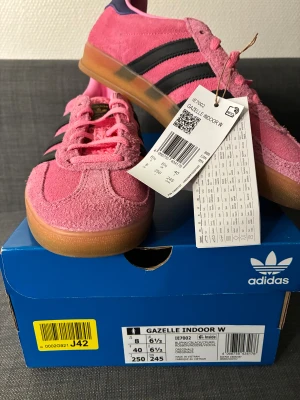 Adidas Gazelle Rosa nya sneakers - Helt nya Adidas Gazelle Indoor sneakers i en snygg rosa färg med svarta ränder och gummisula. Ovandelen är i mjuk mocka och har klassisk snörning. Skon har en rund tå och detaljer i lila vid hälen. Perfekt för dig som vill sticka ut med färg.