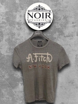 Grå A& Fitch New York t-shirt - Säljer en grå t-shirt från Abercrombie & Fitch med trycket 'A. Fitch New York' på bröstet. T-shirten har korta ärmar, rund halsringning och är tillverkad i mjuk bomull. Perfekt för en avslappnad och stilren look.
