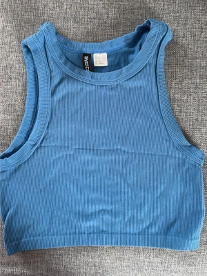 Blå ribbad linnetopp från Divided - Säljer en blå ribbad linnetopp från Divided. Toppen är croppad och ärmlös med rund halsringning. Perfekt för dig som gillar en enkel men trendig stil. Materialet är mjukt och stretchigt, vilket gör den superbekväm.