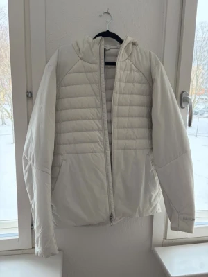 Vit Lulelemon jacka - Säljer en snygg vit pufferjacka med huva och quiltade paneler. Jackan har dragkedja framtill och är tillverkad i ett mjukt, isolerande material som håller dig varm under kalla dagar. Perfekt för vinter och har en modern, clean look.
