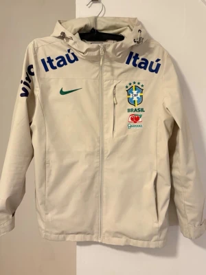 Nike Brasil vindjacka med huva - Stilren beige vindjacka från Nike med Brasilien CBF-logga, grönt swoosh-märke och sponsordetaljer i blått och rött. Jackan har hel dragkedja, huva med dragsko och två sidofickor. Perfekt för dig som gillar sportig streetwear och fotbollsinspirerad stil.
