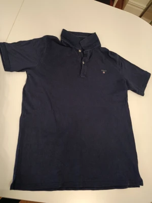 Marinblå pikétröja från Gant - Säljer en klassisk marinblå pikétröja från Gant med korta ärmar och krage. Tröjan har knappar framtill och diskret logotyp på bröstet. Perfekt för en avslappnad och stilren look.