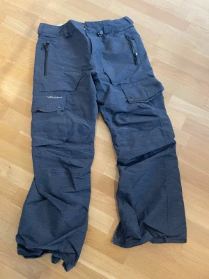 Mörkgrå täckbyxor från Volcom - Säljer ett par mörkgrå täckbyxor från Volcom med flera fickor, dragkedjor och justerbar midja. Byxorna har en avslappnad passform och är perfekta för vinterns alla äventyr. Materialet är slitstarkt och vattenavvisande.