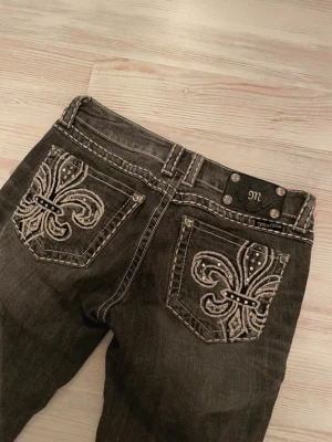 Low waist Miss Me svarta jeans med brodyr - Säljer ett par svarta jeans från Miss Me med snygga broderade fickor bak, dekorerade med vita sömmar och nitar. Jeansen har bootcut-modell och råa kanter nedtill för en cool look. Perfekta för dig som vill sticka ut lite extra. S/XS. W27. Innerbenslängd 80cm, midja tvärs över 36, gren o upp 20, benvidd längst ner 21 ☺️