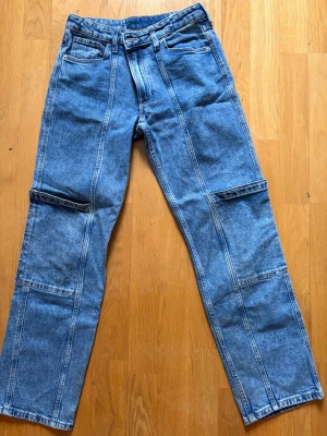 Blå raka jeans från H&M - Säljer ett par blå raka jeans från H&M med låg midja och justerbar midja. Jeansen har tydliga sömmar längs benen och extra detaljer vid knäna för en cool look. Klassisk femficksmodell i slitstark denim, perfekta till vardagsstilen.