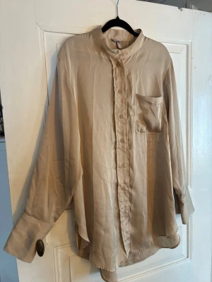 Beige skjorta med ståkrage från H&M - Säljer en beige skjorta från H&M med ståkrage och knappstängning framtill. Skjortan har en bröstficka och långa ärmar med breda manschetter. Materialet är mjukt och har en lätt glans, vilket ger en stilren look.