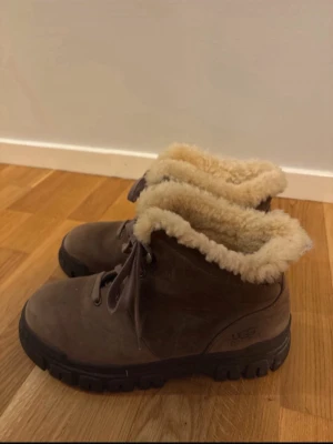 Bruna UGG kängor med fårskinn - Säljer ett par bruna kängor från UGG med grov sula och snörning. Insidan är fodrad med mjukt, ljust fårskinn som syns vid skaftet. Perfekta för kalla dagar och ger en riktigt mysig känsla. Klassisk UGG-logga på sidan.