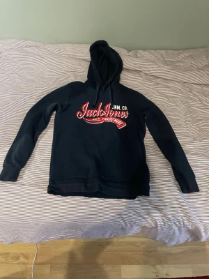 Svart Jack & Jones hoodie med tryck - Svart hoodie från Jack & Jones med stor röd och vit logga på bröstet. Klassisk huva och långa ärmar. Tillverkad i mjukt material som passar perfekt för en avslappnad stil. Snygg och enkel design som funkar till det mesta.