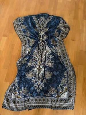 Blå långklänning med orientaliskt mönster - Unik blå långklänning med orientaliskt inspirerat mönster i vitt och gult. Klänningen har snörning i midjan för justerbar passform och korta ärmar. Perfekt för dig som vill sticka ut med en bohemisk och elegant stil.