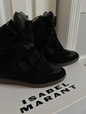 Svarta Isabel Marant sneakers - Snygga svarta sneakers från Isabel Marant med högt skaft och kardborreband. Skorna är gjorda i mocka och har detaljer i läder samt en brun gummisula. Perfekta för dig som vill ha en trendig och sportig look.