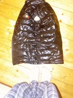 Svart glansig dunväst med patch - Säljer en svart, glansig dunväst med quiltad design och en färgglad patch på bröstet. Västen har dragkedja framtill och sidofickor. Perfekt för lager-på-lager och ger en cool streetwear-look.