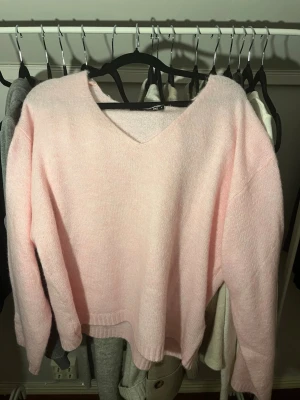 Ljusrosa stickad tröja med V-ringning - Mysig ljusrosa stickad tröja med V-ringning och långa ärmar. Tröjan har en avslappnad passform och ribbade muddar vid ärmslut och nederkant. Perfekt för lager på lager och enkel att matcha med jeans eller kjol.