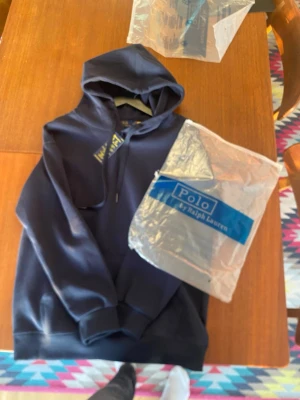 Mörkblå hoodie från Polo Ralph Lauren - Snygg mörkblå hoodie från Polo Ralph Lauren med klassisk huva och dragsko. Hoodien har ribbade muddar vid ärmslut och nederkant samt en enkel, stilren design utan tryck på framsidan. Perfekt för en avslappnad och trendig look.