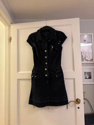 Mörk jeansklänning med knappar - Snygg mörkblå jeansklänning med kort ärm och markerad midja. Klänningen har knappar framtill, krage och flera fickor både fram och bak. Rå kant nertill för en trendig look. Perfekt för dig som gillar en cool och avslappnad stil.