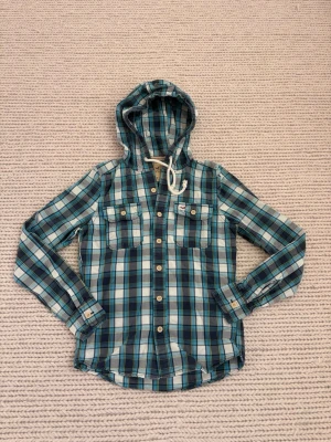 Rutig blå skjorta med huva Hollister - Snygg rutig skjorta från Hollister i blå, vit och grön ton. Skjortan har huva med snörning, två bröstfickor med knappar och långa ärmar. Tillverkad i mjuk bomull, perfekt för en avslappnad stil.