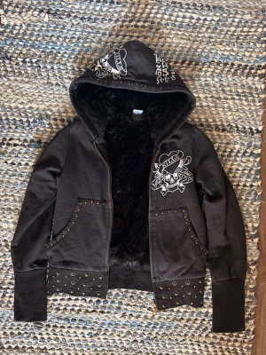 Svart Ed Hardy hoodie med päls - Svart hoodie från Ed Hardy med stora tatueringsinspirerade tryck fram och bak, bland annat dödskalle och hjärta med texten 'Love Kills Slowly'. Mjuk fuskpäls på insidan, nitar och öljetter vid fickor och nederkant. Dragkedja och huva med tryck.
