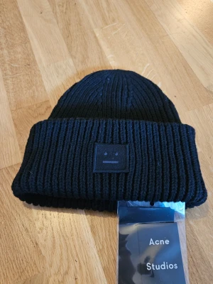 Svart ribbstickad mössa Acne Studios - Säljer en svart ribbstickad mössa från Acne Studios med den ikoniska ansiktsloggan framtill. Mössan är i tjockt, mjuk ull och har en bred uppvikt kant. Perfekt för dig som vill ha en stilren och trendig accessoar till hösten och vintern.