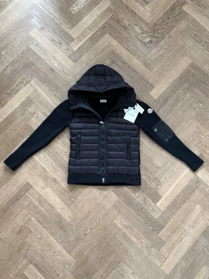 Moncler cardigan - Snygg svart pufferjacka från Moncler med vadderad kropp och stickade ärmar. Jackan har huva, tvåvägsdragkedja och fickor med dragkedja. Moncler-logga på ärmen och praktisk ficka med dragkedja på ena ärmen. Perfekt för kalla vinterdagar. Passar 180-185