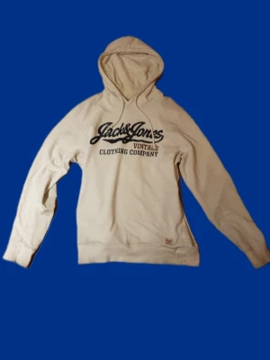 hoodie Jack & Jones - Snygg vit hoodie från Jack & Jones med trycket 'Vintage Clothing Company' i svart på bröstet. Tillverkad i mjuk bomull och har klassisk huva med dragsko. Perfekt för en avslappnad och trendig stil.