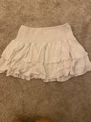 Vit volangkjol med resårmidja - Supersöt vit kortkjol med resår i midjan och flera lager volanger. Kjolen är gjord i ett lätt tyg som känns skönt mot huden. 