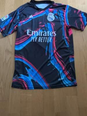Real Madrid Bellingham matchtröja - Snygg Real Madrid matchtröja från Adidas med Bellingham och nummer 5 på ryggen. Tröjan är svart med färgstarka blå och rosa mönster, korta ärmar och klubbmärke samt sponsortryck på bröstet. Tillverkad i lätt och ventilerande funktionsmaterial.