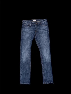 Jack & Jones - Säljer ett par klassiska blå jeans från Jack n Jones med smal passform och snygg slitning. Jeansen har fem fickor, dragkedjegylf och detaljerade sömmar på bakfickorna. Tillverkade i slitstarkt denim med en cool tvättad look.
