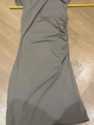 Grå långklänning med rynkade detaljer - Snygg grå långklänning med rynkade detaljer på sidan. Klänningen har en enkel och stilren design med mjukt, stretchigt material som ger en smickrande passform. Perfekt för dig som gillar minimalistisk stil.