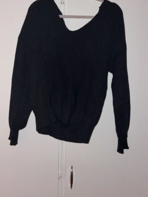 Svart stickad tröja med rosett  - Säljer en svart stickad tröja med v-ringning och en snygg satinrosett i nacken. Tröjan har ballongärmar och ribbade muddar vid ärmslut och midja. Perfekt för dig som gillar en enkel men ändå unik look.