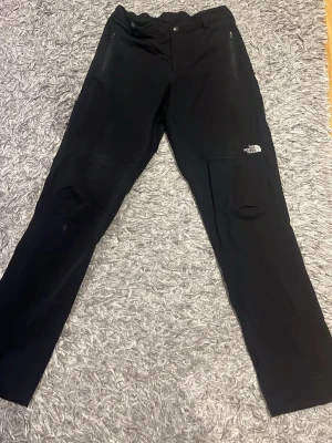 Svarta cargobyxor från The North Face - Snygga svarta cargobyxor från The North Face med raka ben och dragkedjeförsedda fickor framtill. Byxorna har en diskret logga på låret och är tillverkade i ett slitstarkt, funktionellt material som passar perfekt för en aktiv livsstil.