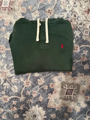 Grön hoodie från Polo Ralph Lauren - Mörkgrön vintage hoodie från Polo Ralph Lauren med klassisk röd logga broderad på bröstet. Hoodien har vita dragsnören, känguruficka och ribbade muddar. Tillverkad i mjukt bomullsmaterial för en skön och avslappnad stil. Det är lite slitningar men syns inte så värst på den samt få fläckar. Det är M/M men är stor i storlek.