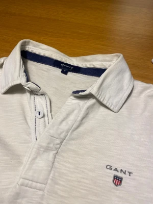 Vit henley från Gant med brodyr - Vit henley från Gant med klassisk krage, knappslå och broderad logga på bröstet. Tillverkad i mjuk bomull och har en stilren look med diskreta detaljer. Perfekt för dig som gillar enkel men snygg stil.