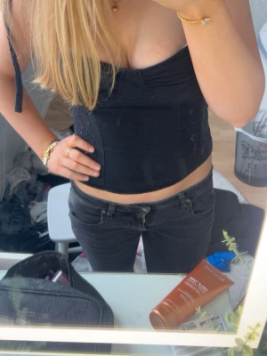 Svart topp med knytband - Svart topp med knytbara axelband och dekorativa broderade detaljer på sidorna. Toppen har en dragkedja i ryggen och är croppad i modellen. Perfekt för dig som vill ha en trendig och stilren look. Toppen är ifrån Zara. Toppen har ett litet hål precis vid dragkedjan, som du ser på bild 5. Påverkar dock inte själva dragkedjan.