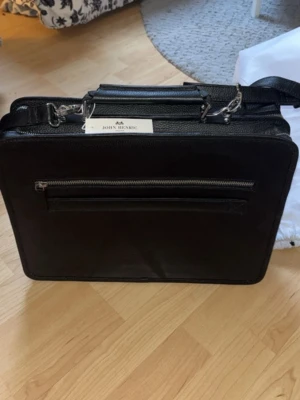 Svart portfölj från John Henric - Snygg svart portfölj i läder från John Henric med både handtag och axelrem. Väskan har flera fack, bland annat en stor ficka med dragkedja på framsidan och två mindre fickor på baksidan. Perfekt för laptop och dokument. Stilig och praktisk design.