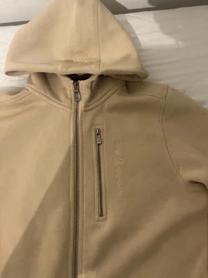 Beige hoodie från Sail Racing - Snygg beige hoodie från Sail Racing med dragkedja framtill, bröstficka med zip och broderad logga. Hoodien har en stilren design, huva och är tillverkad i mjukt material som känns skönt mot huden. Perfekt för en avslappnad och sportig look.