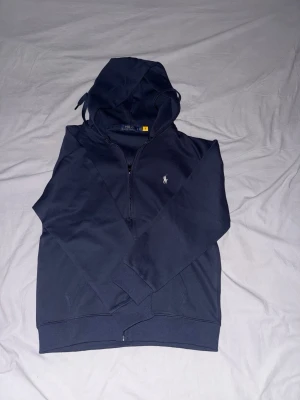 Mörkblå hoodie från Polo Ralph Lauren - Snygg mörkblå hoodie från Polo Ralph Lauren med dragkedja framtill och klassisk huva med dragsko. Ikoniska loggan broderad på bröstet. Tillverkad i mjukt bomullsmaterial och har ribbade muddar vid ärmslut och nederkant. Perfekt för en chill och stilren look.