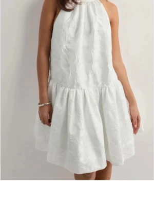 By Malin Flora minidress stl s ny! - Supersöt vit kort klänning med volangkant och strukturmönster i tyget. Klänningen har ärmlös design och halterneck-ringning som ger en fräsch och modern look. Perfekt för studenten. Helt ny med lapparna  kvar! Köpte för 2700.