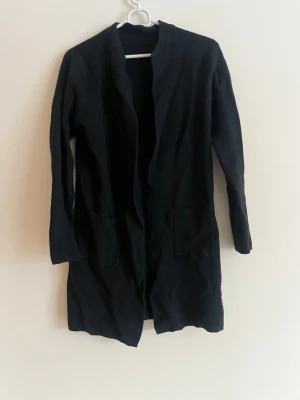 Svart lång cardigan med fickor - En svart lång cardiganjacka med öppen front och två stora fickor framtill. Jackan har lång ärm och rak passform, perfekt att slänga över en t-shirt eller hoodie. Materialet ser ut att vara mjukt och stickat, vilket gör den extra skön.