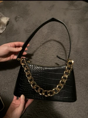 Svart axelväska med guldkedja - Snygg svart handväska i croco-mönstrat konstläder med en chunky guldfärgad kedja framtill. Väskan har en enkel axelrem och stängs med dragkedja. Perfekt accessoar för att lyfta din outfit med en trendig touch. Den har ett litet veck på axelbandet.