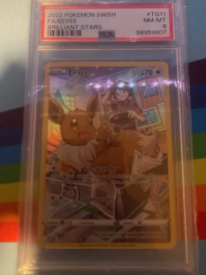 Pokémonkort Eevee Brilliant Stars PSA - Säljer ett samlarkort från Pokémon, Eevee FA/Brilliant Stars 2022, graderat av PSA med betyg 8. Kortet är inneslutet i ett genomskinligt plastskydd och har en färgglad, holografisk design med Eevee och en tränare i bakgrunden. Kan också ta byten men vill helst sälja