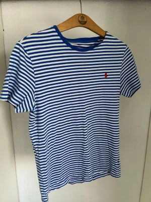Randig t-shirt från Ralph Lauren - Snygg t-shirt från Ralph Lauren med blå och vita ränder och en liten röd logga broderad på bröstet. Klassisk passform med rund halsringning och korta ärmar. Tillverkad i mjuk bomull som känns skön mot huden.