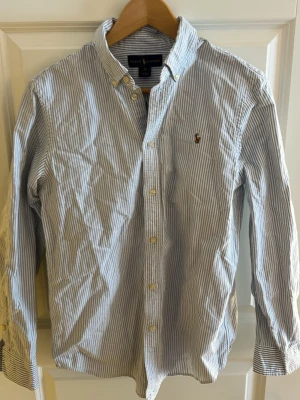 Randig skjorta från Ralph Lauren - Klassisk blåvit randig skjorta från Ralph Lauren med button-down krage och broderad logga på bröstet. Skjortan har långa ärmar och knäppning framtill. 