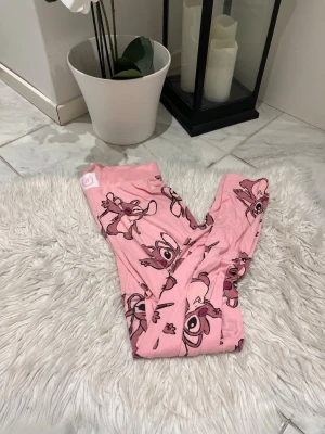 Rosa pyjamasbyxor med Stitch-motiv - Säljer ett par rosa pyjamasbyxor från H&M med gulligt Stitch-tryck över hela tyget. Byxorna är mjuka och har resår i midjan för extra komfort. Perfekta för mysiga hemmakvällar eller när du bara vill chilla. ( gått isär bakifrån men går att sy ihop ) 