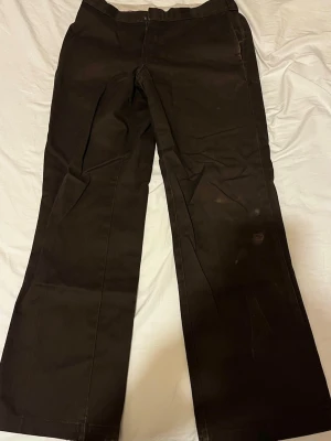 Bruna chinos från Dickies - Säljer ett par bruna chinos från Dickies med raka ben och klassisk passform. Byxorna har bälteshällor, fickor fram och bak samt är tillverkade i ett slitstarkt bomullsmaterial. Perfekta för en avslappnad och stilren look.